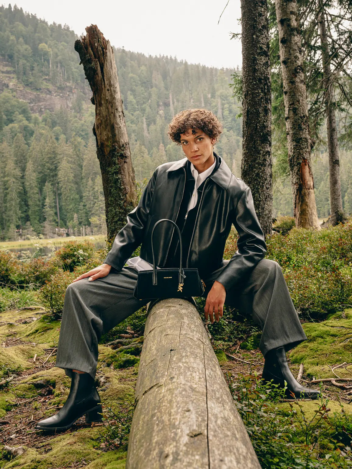 Frau sitzt im Wald in schwarzer Lederjacke, weißem Hemd und grauer Wide-Leg-Hose, schwarze Michael-Kors-Tasche auf dem Bein abgestellt – Herbst/Winter-Outfit 2026 von FREY Mode