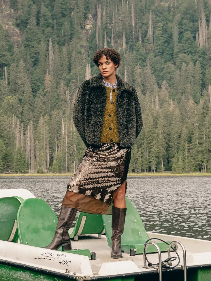 Frau auf Tretboot trägt Plüschjacke über Steppweste und Streifenhemd, Pailletten-Rock und hohe Lederstiefel – Herbst-Outfit 2026 von FREY Mode