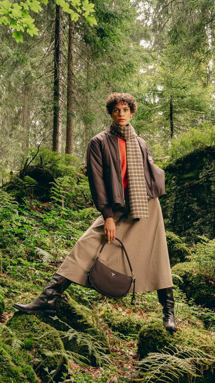 Frau im Wald in Karo-Look: brauner Wendejacke, orangenem Pullover, kariertem Schal, Karo-Midirock und Lederstiefeln, hält braune Hobo-Tasche in der Hand – Herbst-Outfit 2026 von FREY Mode