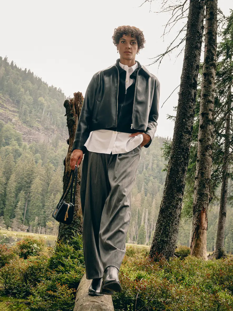 Frau im Wald trägt schwarze Lederjacke über weißem Hemd, graue Wide-Leg-Hose und schwarze Stiefeletten, hält schwarze Schultertasche – Herbst-Outfit 2026 von FREY Mode