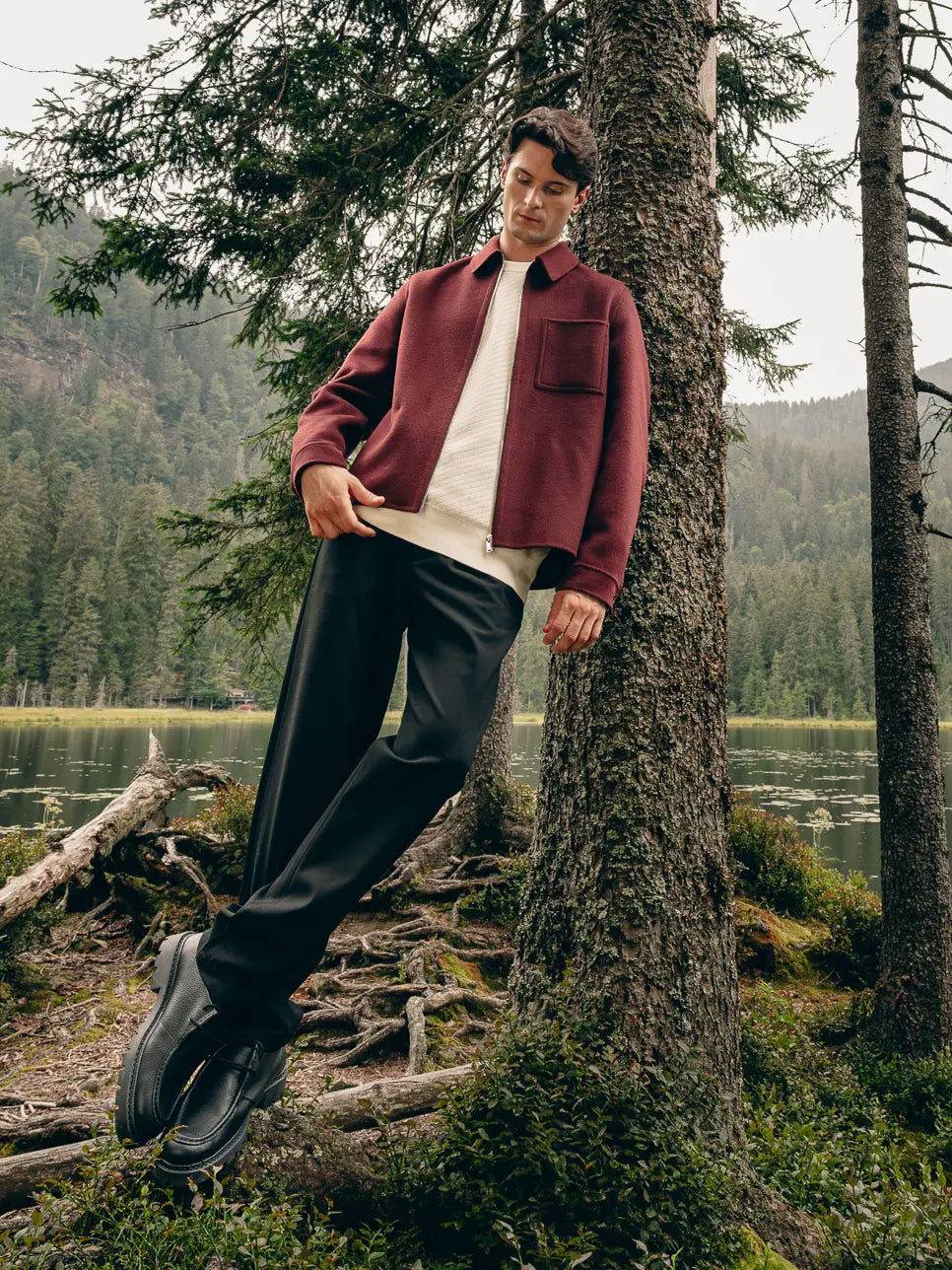 Mann lehnt im Wald an Baum, trägt braune Hemdjacke über cremefarbenem Pullover mit Bordeaux-T-Shirt und schwarzer Hose – Herbst-Outfit 2026 von FREY Mode
