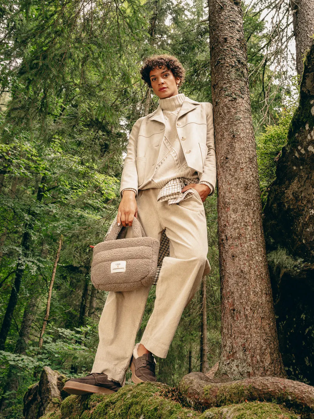Frau im Wald trägt beige Boxy-Jacke über Pullover, kariertes Hemd um die Hüfte gebunden und beige Hose, hält beige Teddy-Tasche – Herbst-Outfit 2026 von FREY Mode