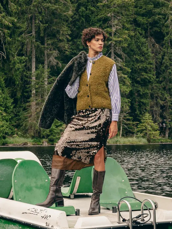 Frau steht auf Tretboot in Layering-Look aus Plüschjacke, Weste, Streifenhemd und Pailletten-Rock mit hohen Stiefeln – Herbst/Winter-Outfit 2026 von FREY Mode