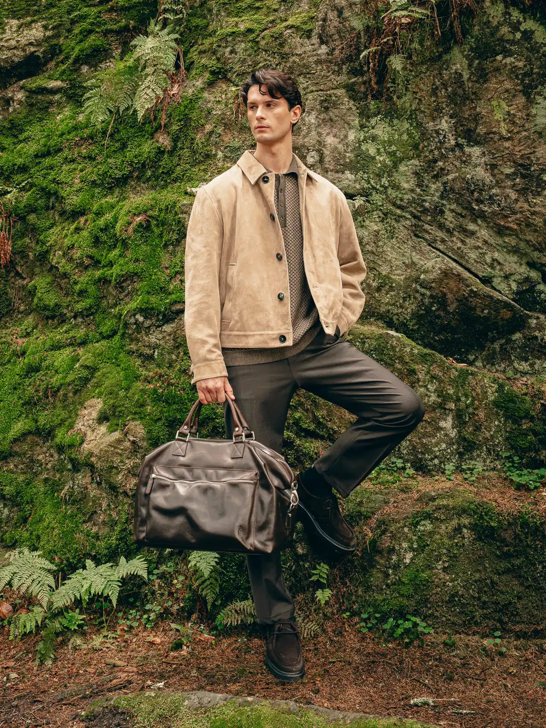 Mann steht im Wald in Braunton-Look aus Veloursleder-Jacke und Wollhose mit brauner Reisetasche und Loafern – Herbst/Winter-Outfit 2026 von FREY Mode