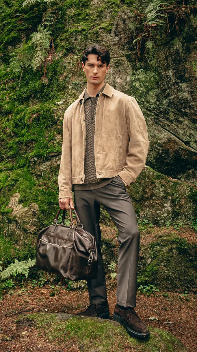 Mann trägt beige Veloursleder-Jacke über gemustertem Pullover und graue Hose, hält braune Reisetasche – Herbst-Outfit 2026 von FREY Mode