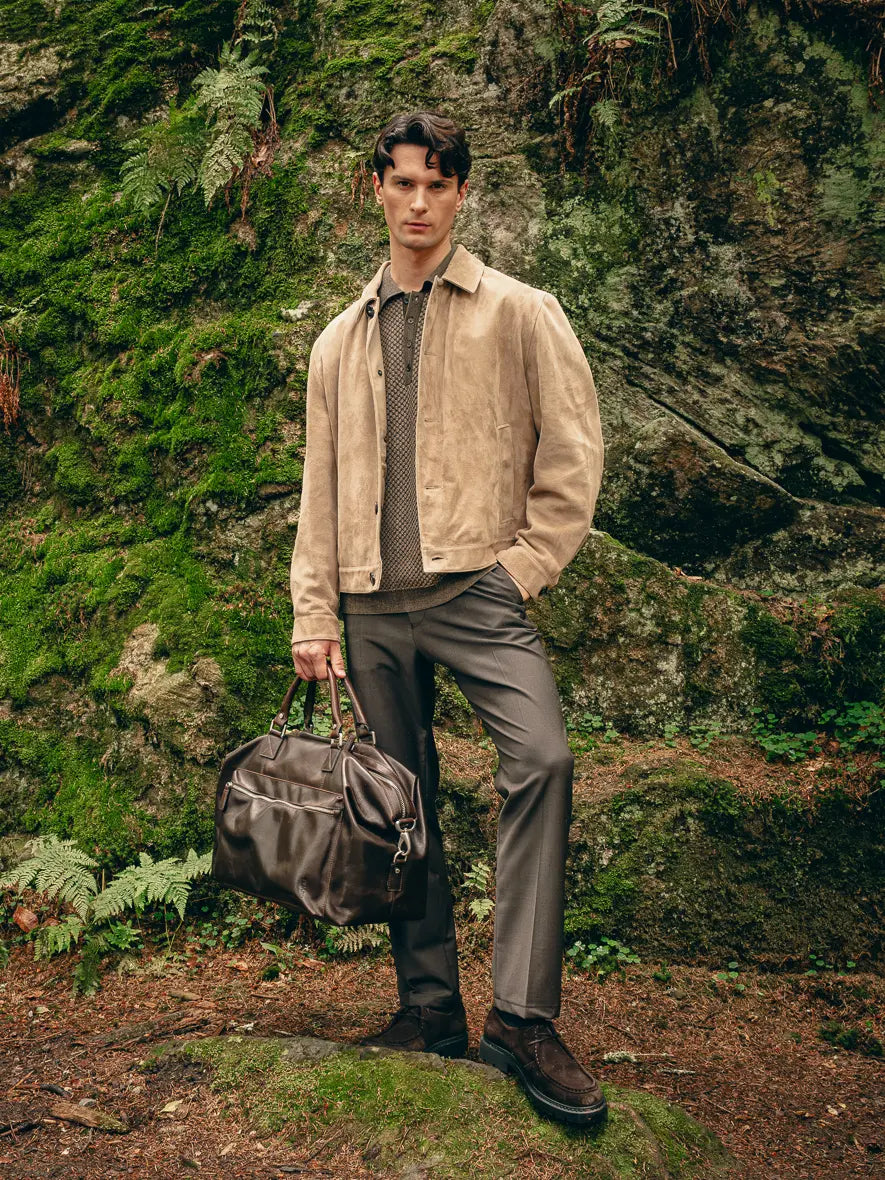Mann trägt beige Veloursleder-Jacke über gemustertem Pullover und graue Hose, hält braune Reisetasche – Herbst-Outfit 2026 von FREY Mode