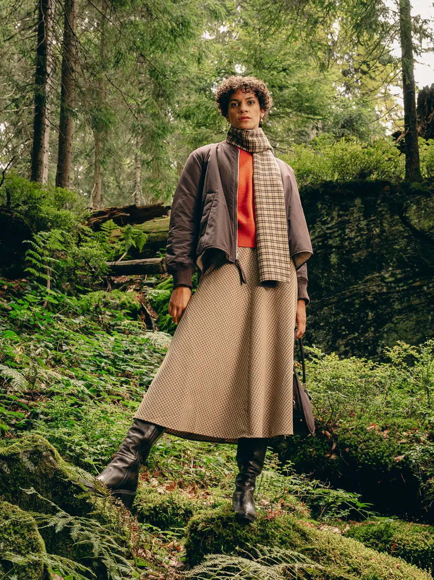 Frau steht im Wald in Karo-Layering-Look aus brauner Wendejacke, orangenem Pullover, kariertem Schal und Karo-Midirock mit Lederstiefeln – Herbst/Winter-Outfit 2026 von FREY Mode