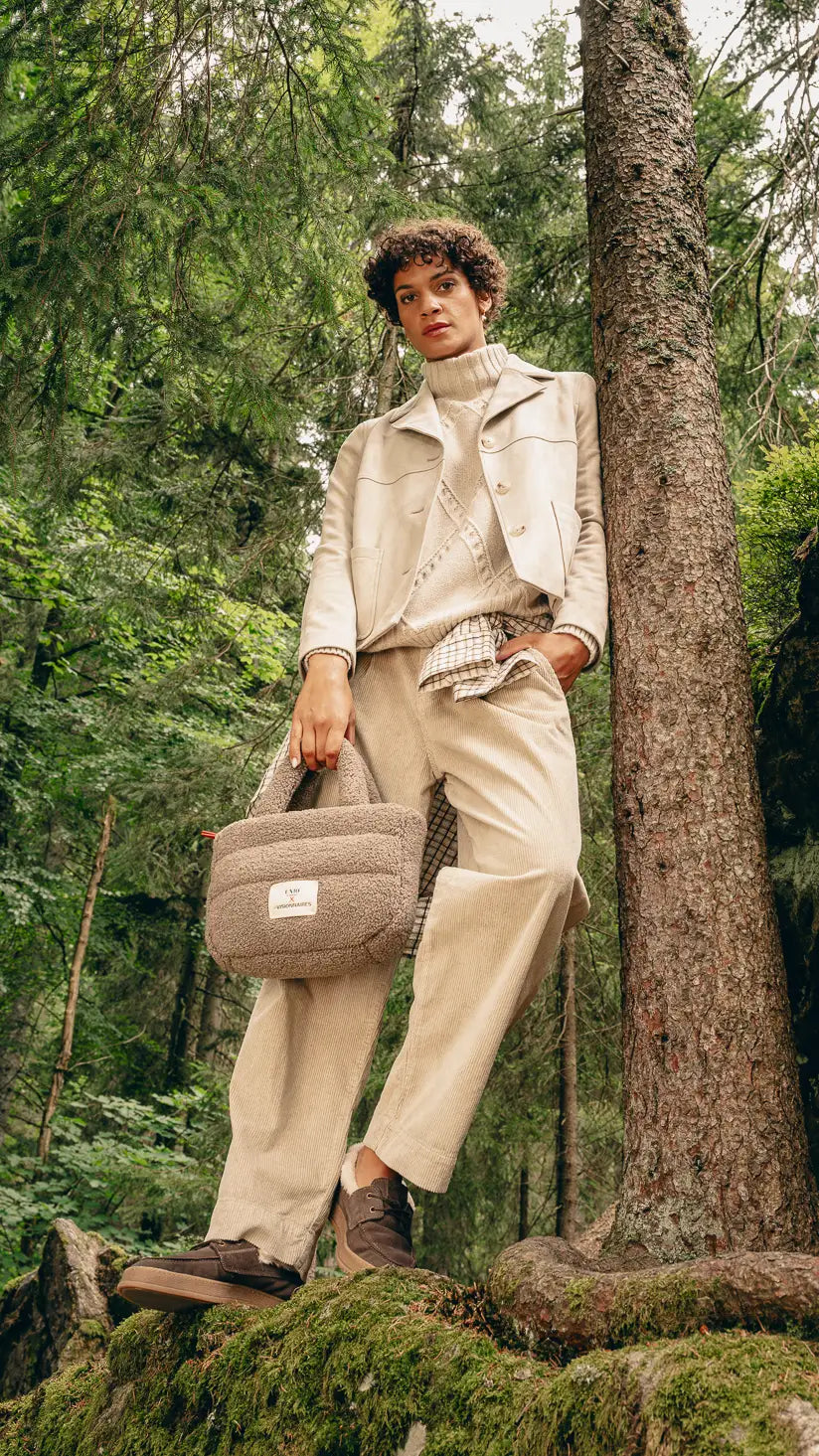 Frau im Wald trägt beige Boxy-Jacke über Pullover, kariertes Hemd um die Hüfte gebunden und beige Hose, hält beige Teddy-Tasche – Herbst-Outfit 2026 von FREY Mode