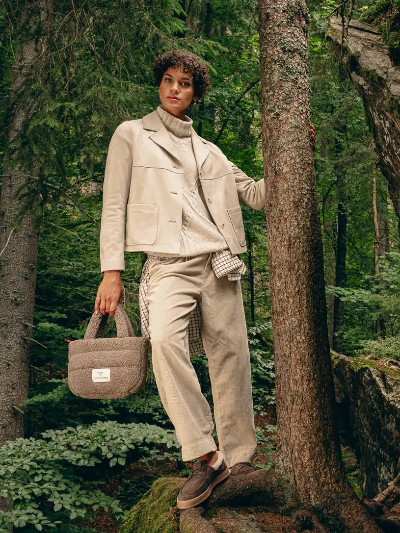Frau lehnt im Wald an einem Baum, trägt beige Boxy-Jacke, Loose-Fit-Pullover und Hose mit kariertem Hemd um die Hüfte gebunden – Herbst/Winter-Outfit 2026 von FREY Mode