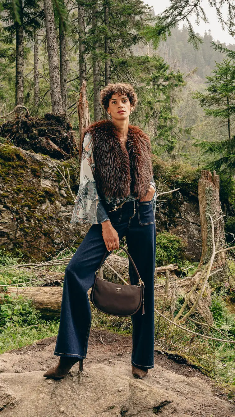 Frau im Wald trägt braune Kunstfellweste, Volant-Bluse und dunkelblaue Wide-Leg-Jeans – Herbst-Outfit 2026 von FREY Mode