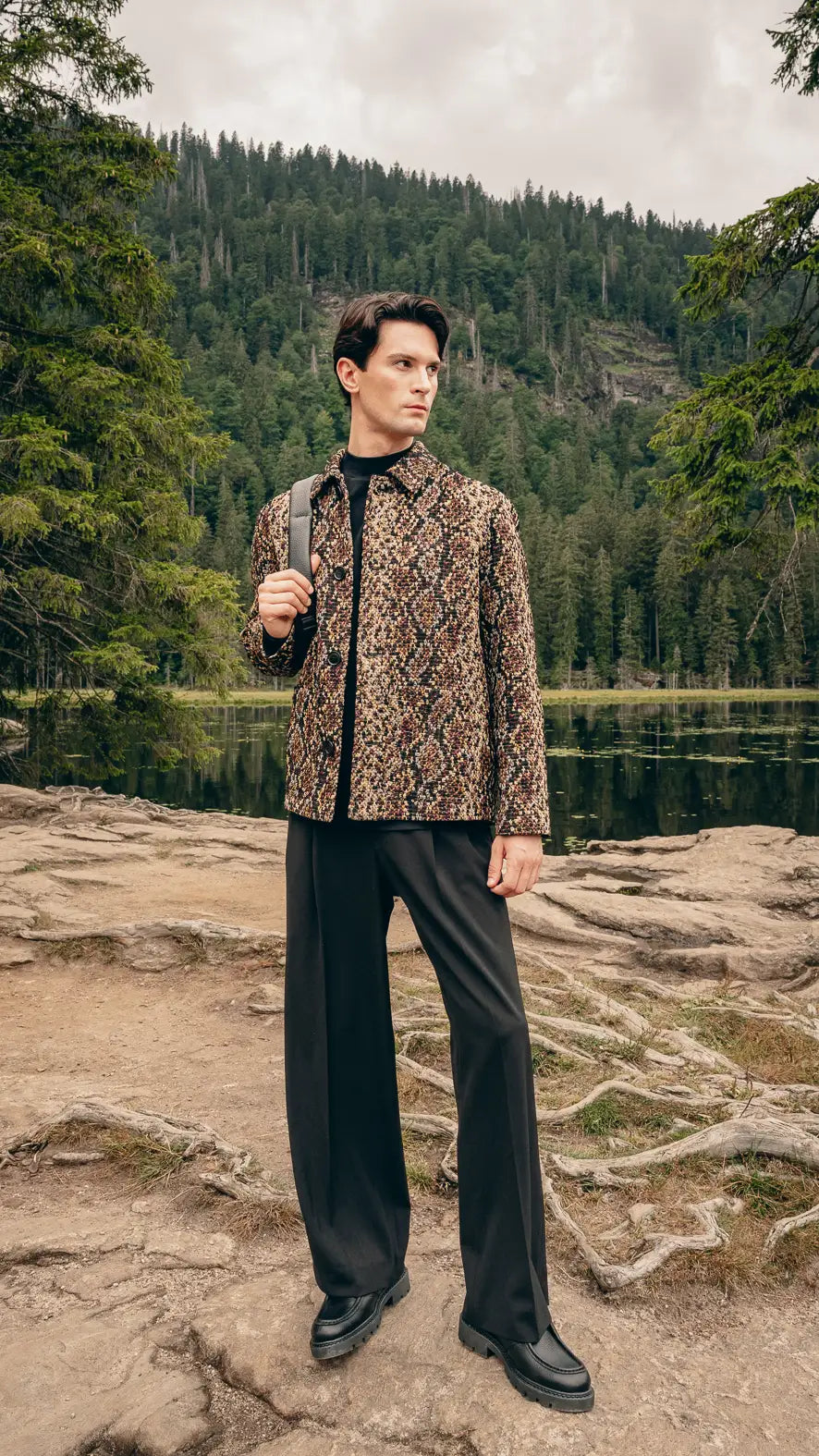 Mann steht auf Felsen am See in strukturierter Jacke und extrem weiter schwarzer Hose, hält schwarzen Leder-Rucksack – Volumen-Trend Herbst/Winter 2026 von FREY Mode