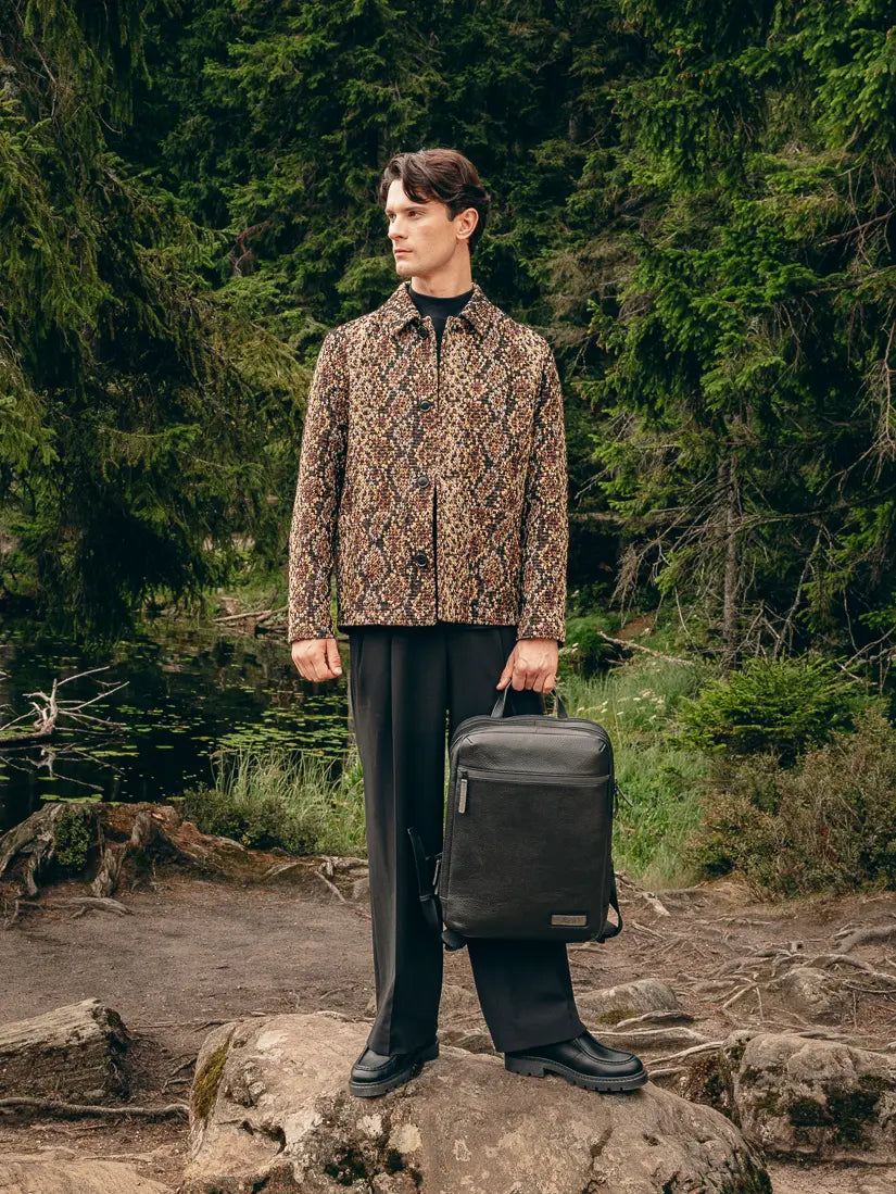 Mann steht auf Felsen am See in strukturierter Jacke und extrem weiter schwarzer Hose, hält schwarzen Leder-Rucksack – Volumen-Trend Herbst/Winter 2026 von FREY Mode