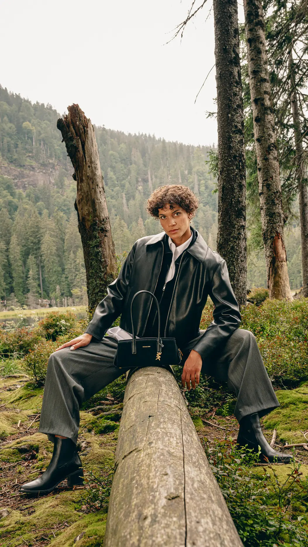 Frau sitzt im Wald in schwarzer Lederjacke, weißem Hemd und grauer Wide-Leg-Hose, schwarze Michael-Kors-Tasche auf dem Bein abgestellt – Herbst/Winter-Outfit 2026 von FREY Mode