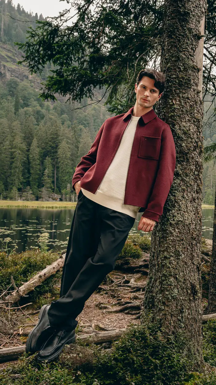 Mann lehnt im Wald an Baum, trägt braune Hemdjacke über cremefarbenem Pullover mit Bordeaux-T-Shirt und schwarzer Hose – Herbst-Outfit 2026 von FREY Mode