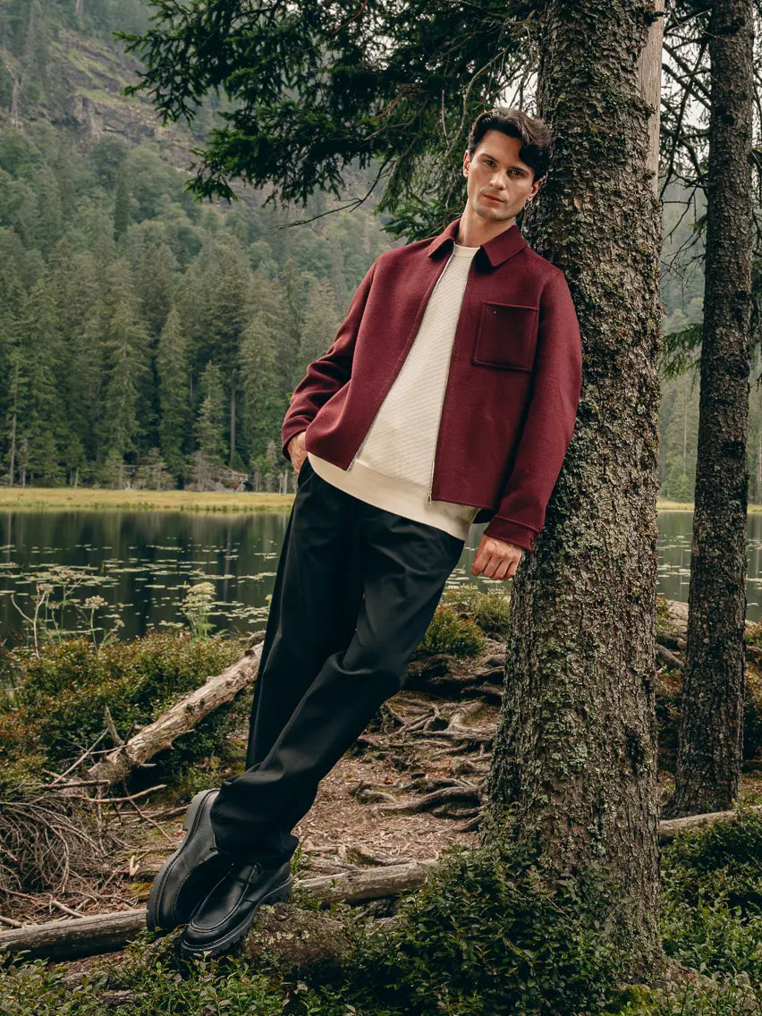 Mann lehnt im Wald an Baum, trägt braune Hemdjacke über cremefarbenem Pullover mit Bordeaux-T-Shirt und schwarzer Hose – Herbst-Outfit 2026 von FREY Mode